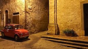 A narrow street with a vintage red car parked on the left, a stone wall on the right, and a small potted plant at the corner.