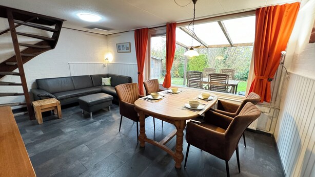 Een gezellige woonkamer met een houten eettafel, beklede bruine stoelen, een zwarte leren bank en een houten trap.
