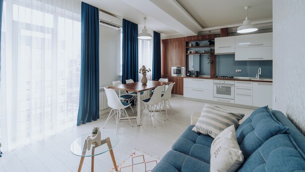 A modern living room with a blue sofa, a glass coffee table, and a dining area with a wooden table and white chairs.
