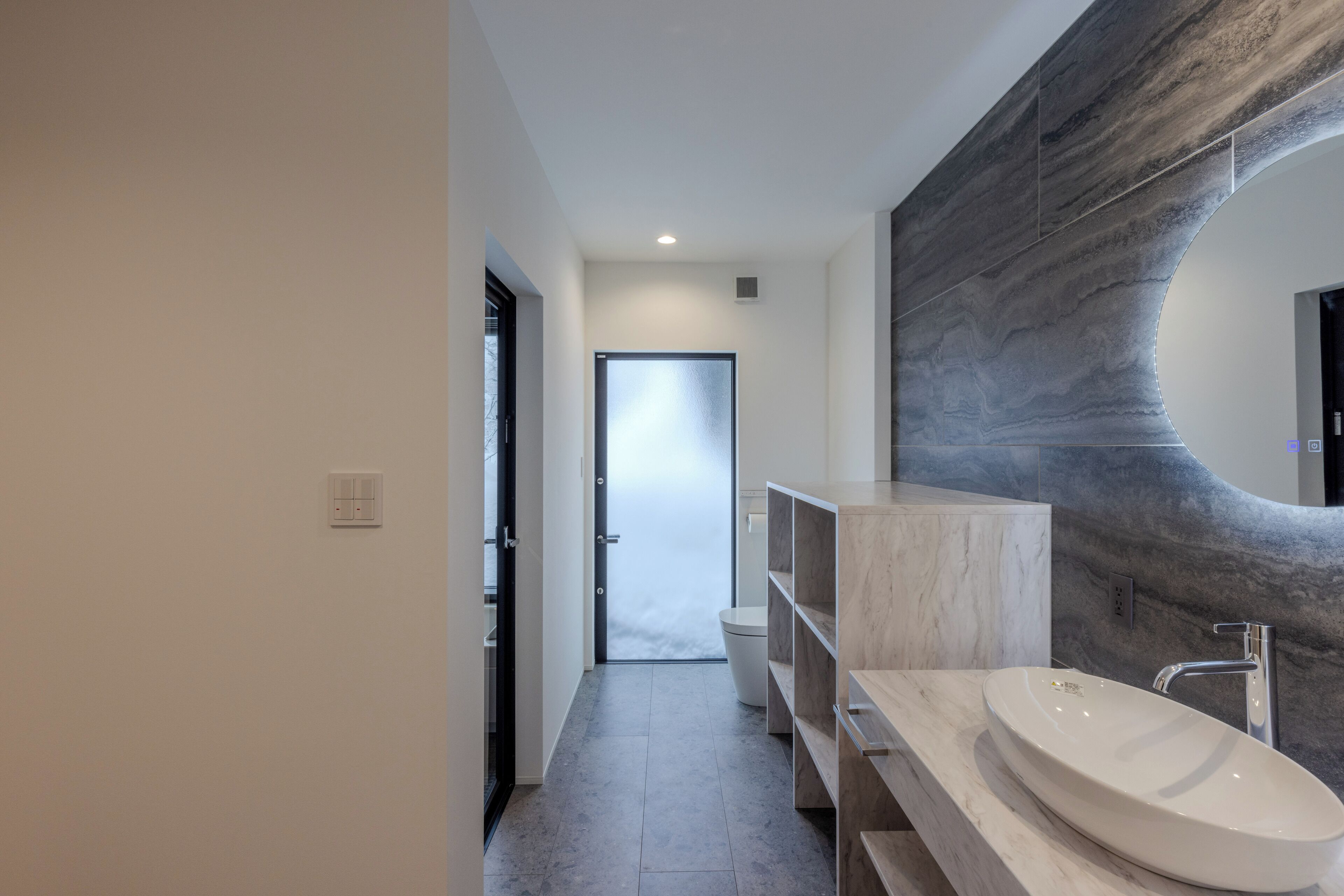 A modern bathroom with a large oval sink, a curved marble wall, and a glass door leading to a toilet.