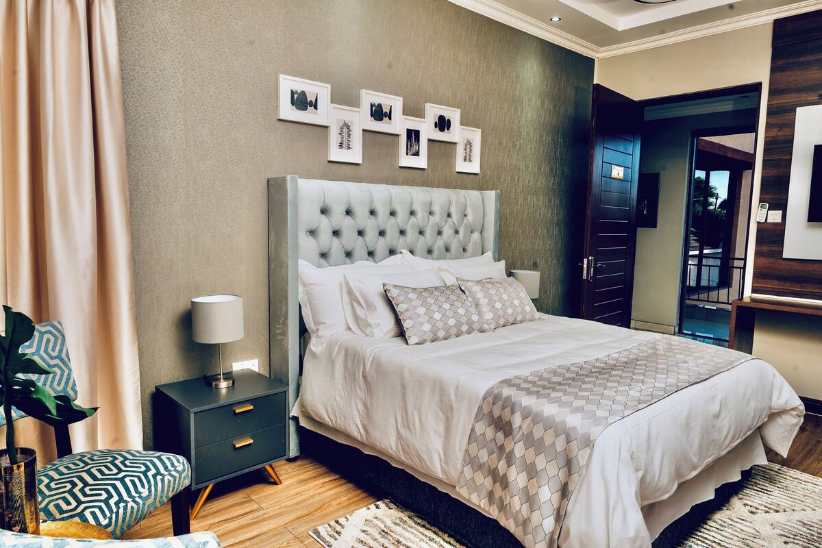 A modern bedroom with a tufted headboard, a bed with a patterned bedspread, a bedside table with a lamp, and a wall with framed artwork.
