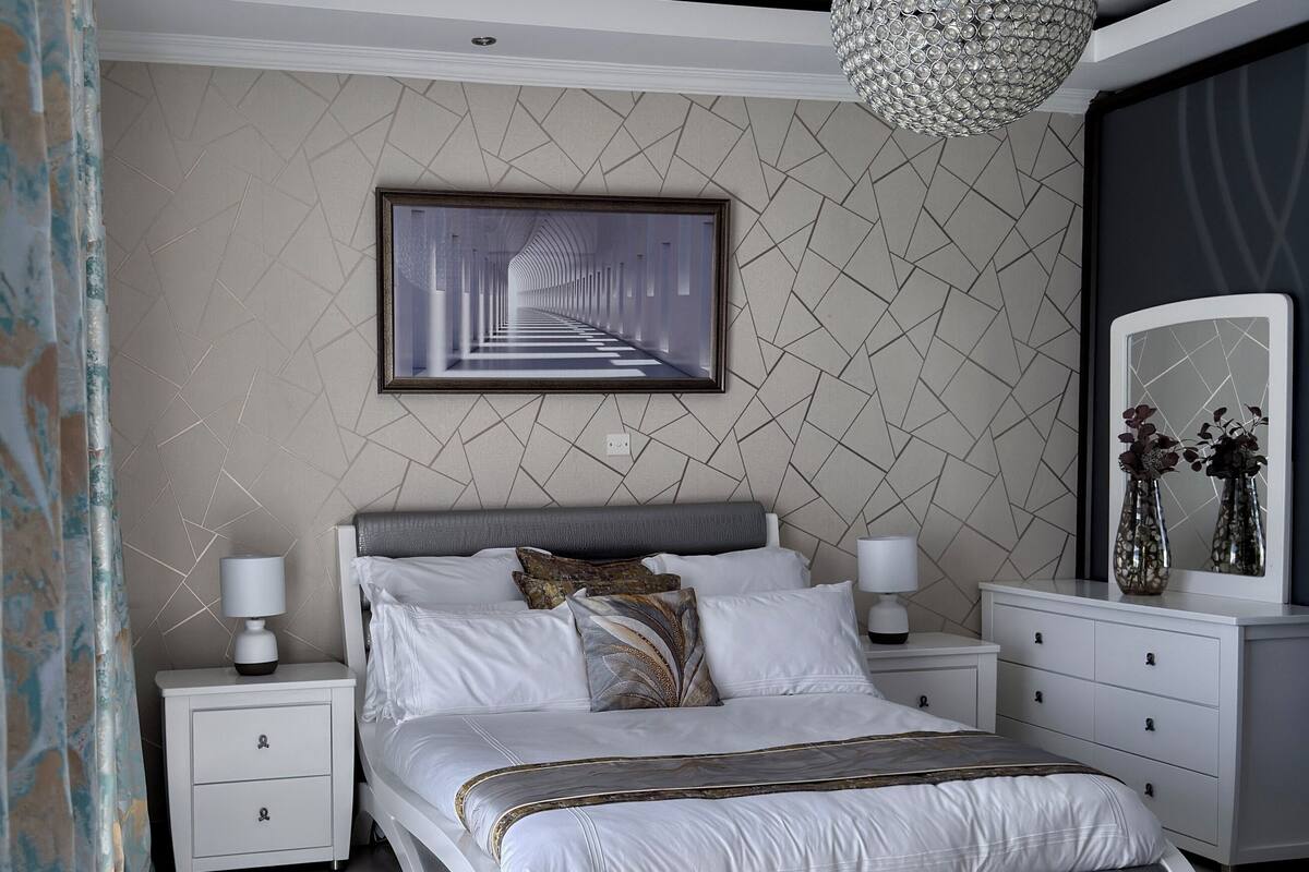 A modern bedroom with a large bed, bedside tables, a chandelier, and a geometric-patterned wall.