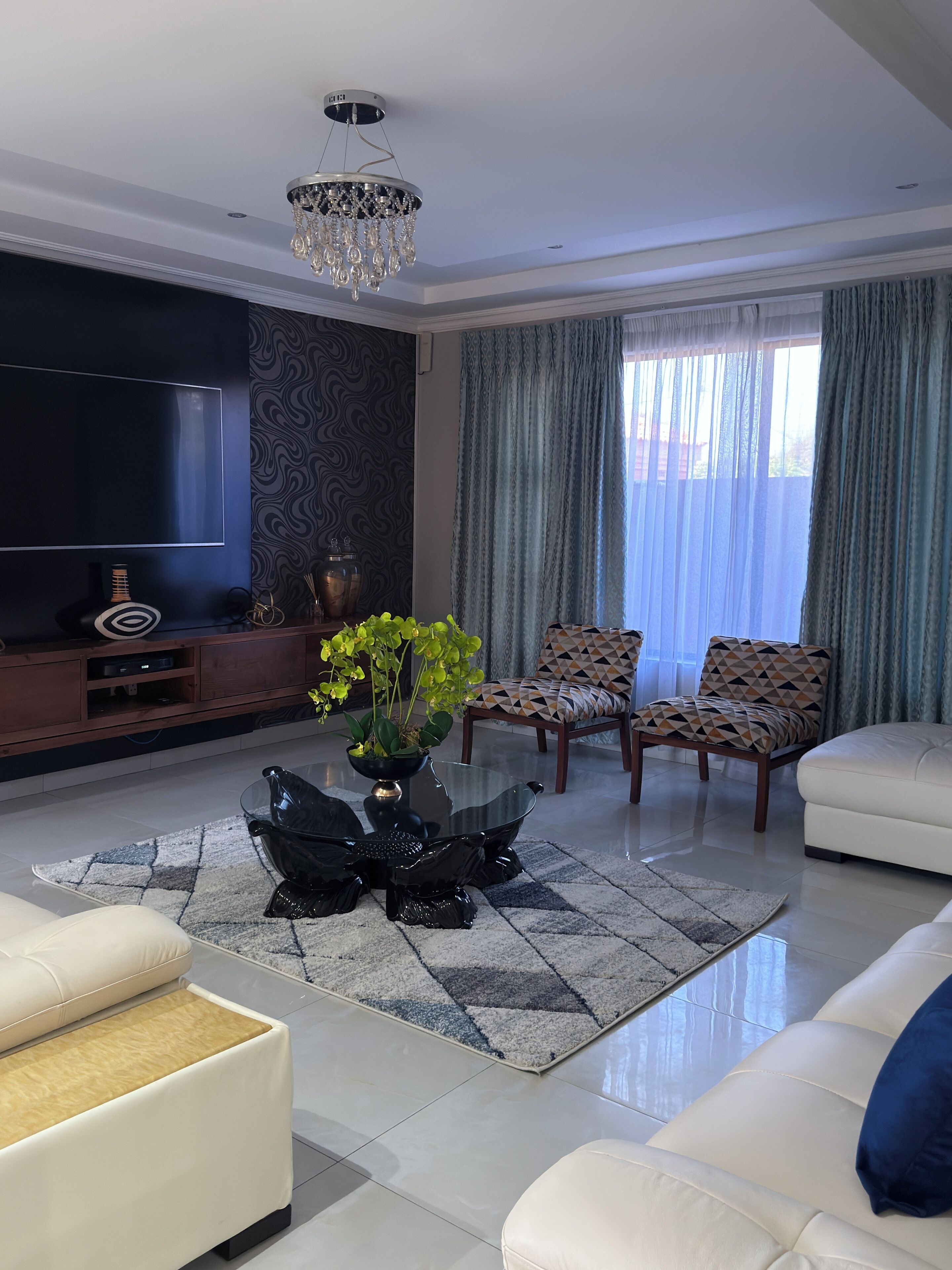 A modern living room with a flat-screen TV, a chandelier, patterned curtains, and a coffee table with a plant.