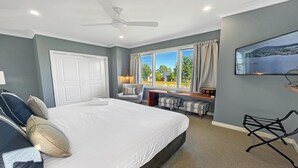 Quarto de hotel com uma cama grande, escrivaninha, cadeira, ventilador de teto e janela com vista para área verde.