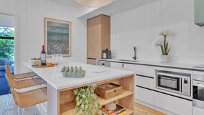 A modern kitchen with white cabinetry, a wooden accent wall, a large island with seating, and a view of greenery outside. - Birdsong Beach Retreat (Skennars Head)