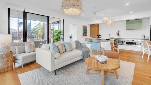 A modern living room with a sofa, coffee table, and chairs, a kitchen area with a dining table, and large windows offering a view of the outdoors. - Birdsong Beach Retreat (Skennars Head)