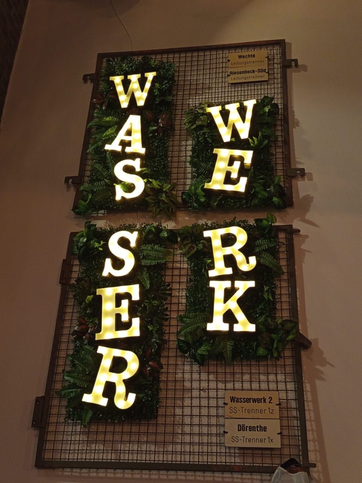 A wall with illuminated letters spelling "WAS WE RK" surrounded by greenery. Below, there are signs indicating "Wasserwerk 2 SS-Trenner 1z" and "Dörrenthe SS-Trenner 1x".