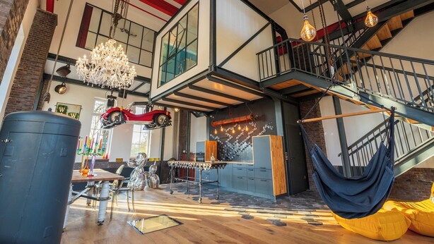 Un loft spacieux aux murs en briques apparentes, doté d’une mezzanine, d’un luminaire suspendu et d’une voiture rouge d’époque accrochée au plafond.