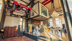 Un loft spacieux avec une voiture de sport rouge suspendue au plafond, un coin repas agrémenté d’un lustre, et un escalier menant à un niveau supérieur.