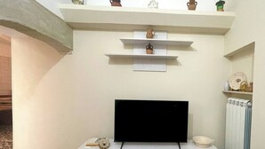Una sala de estar con televisión de pantalla plana, estanterías blancas con objetos decorativos, suelo de mármol y un radiador.