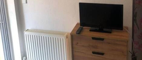 Una habitación con una televisión sobre un mueble de madera, un radiador y una estantería con objetos decorativos.