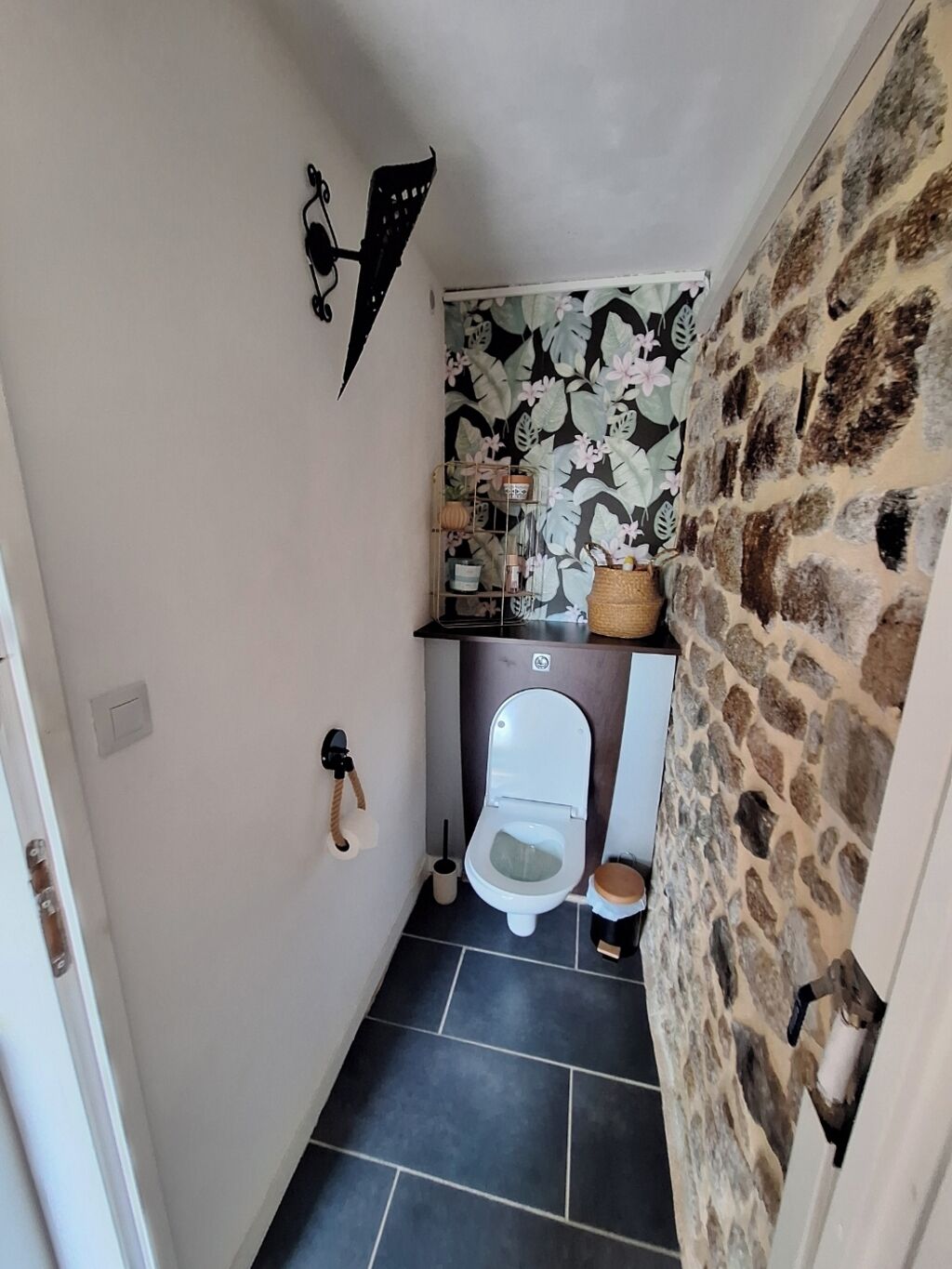 A narrow bathroom with a toilet, a small shelf with toiletries, and a decorative wall with a leaf pattern.