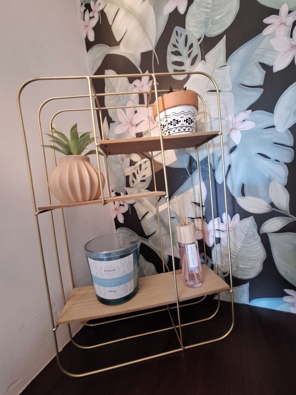 A three-tiered metal shelf with a potted plant, a candle, and a decorative item against a floral wallpaper background.