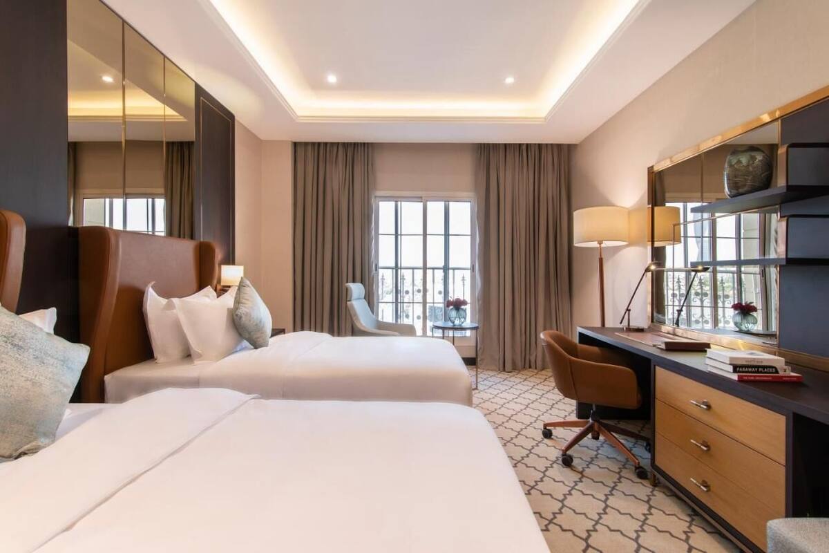 Habitación de hotel con dos camas, un escritorio, una silla, una mesita pequeña y un ventanal grande.