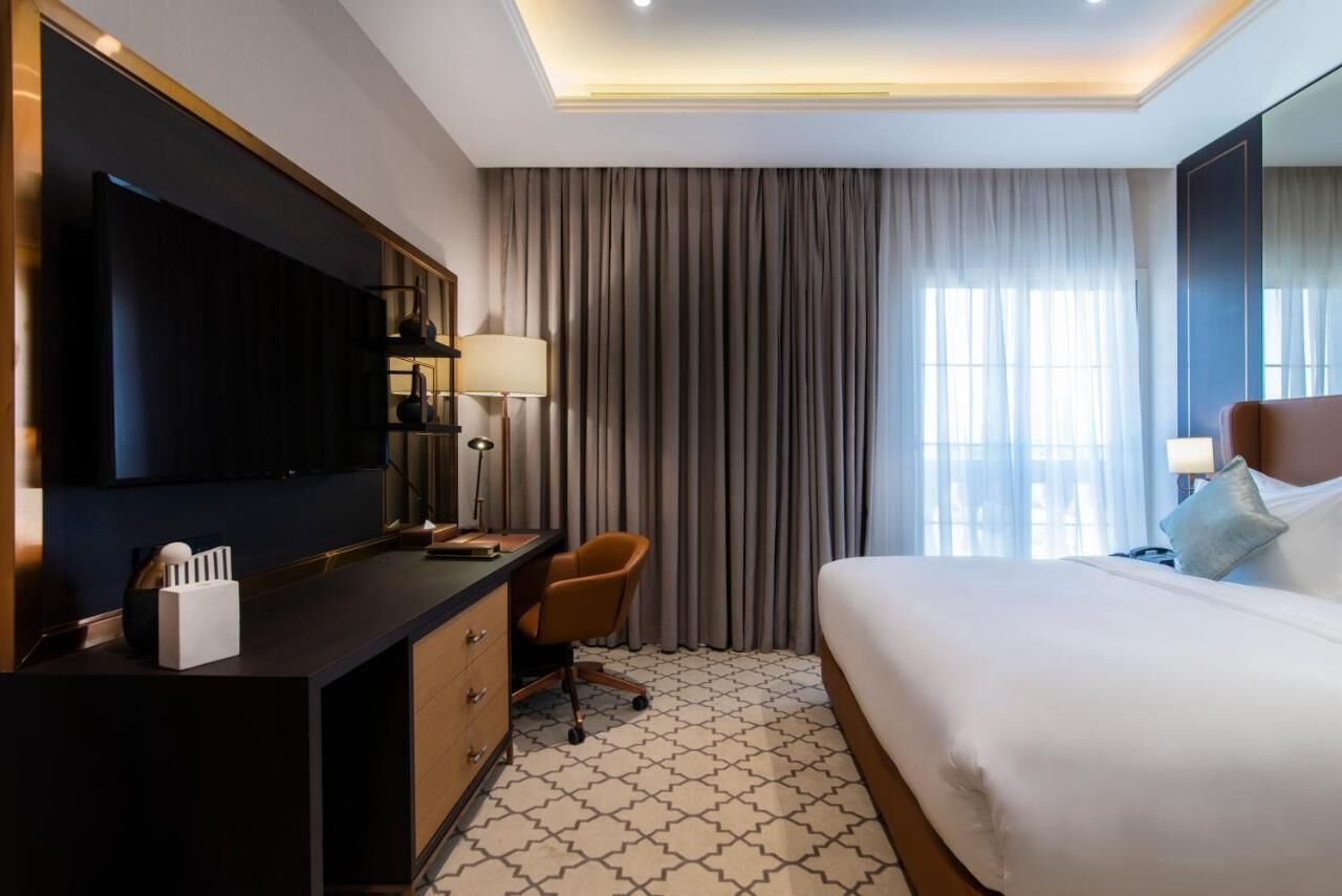 A hotel room with a bed, a desk with a chair, a TV, and a large window with curtains.