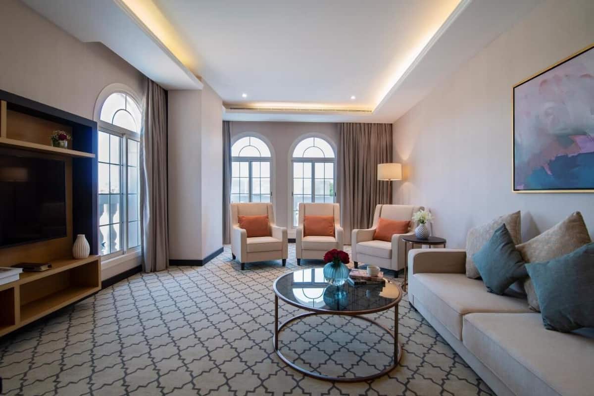Una sala de estar moderna con un sofá, dos sillones, una mesa de centro y un televisor. Hay ventanas amplias con cortinas, un suelo con diseño y un cuadro en la pared.