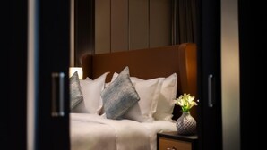Un letto rifatto con lenzuola bianche e due cuscini, un comodino con un vaso di fiori, e un armadio scuro sullo sfondo.