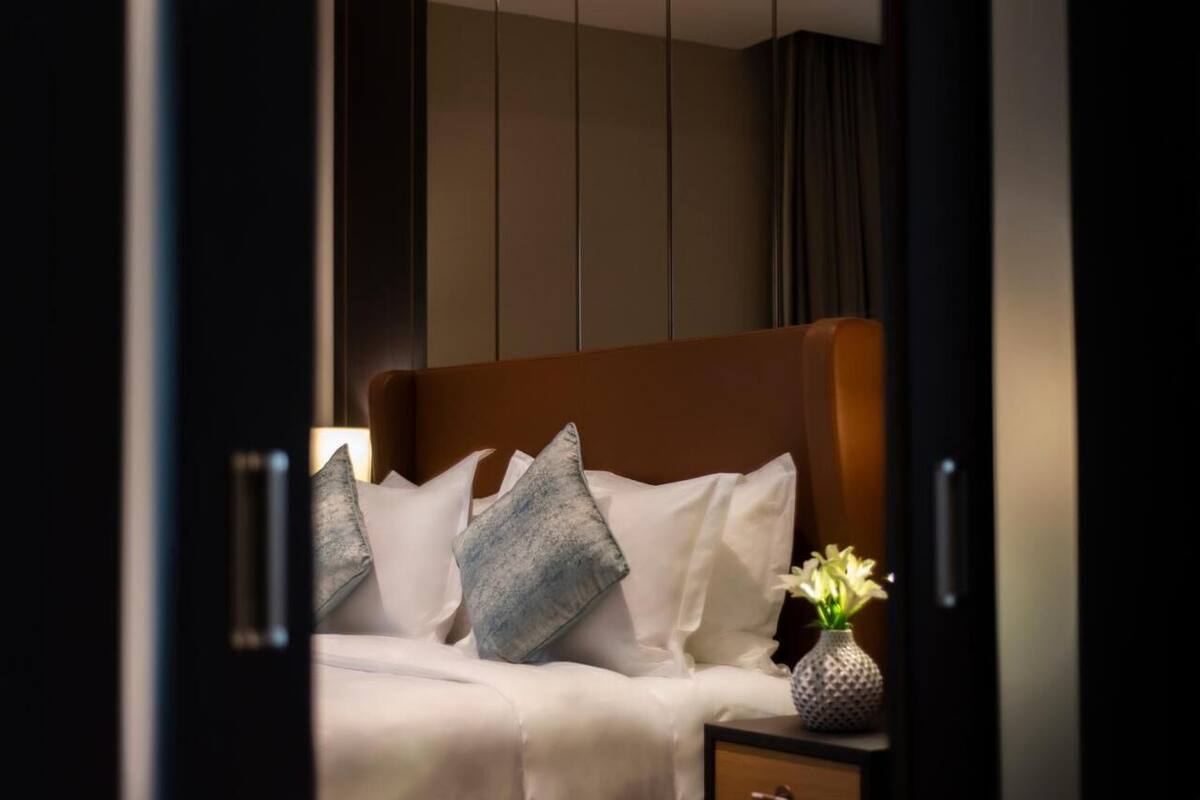 Una cama bien hecha con sábanas blancas y dos almohadas, una mesita de noche con un florero, y un armario oscuro al fondo.