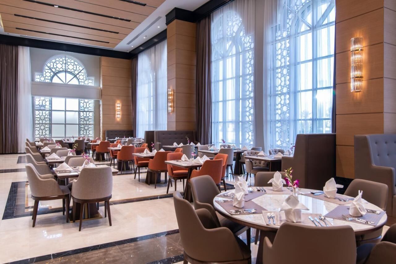 Un vaste espace de restauration dans l’hôtel, avec de nombreuses tables et chaises, de grandes fenêtres ornées de motifs décoratifs et un éclairage mural.