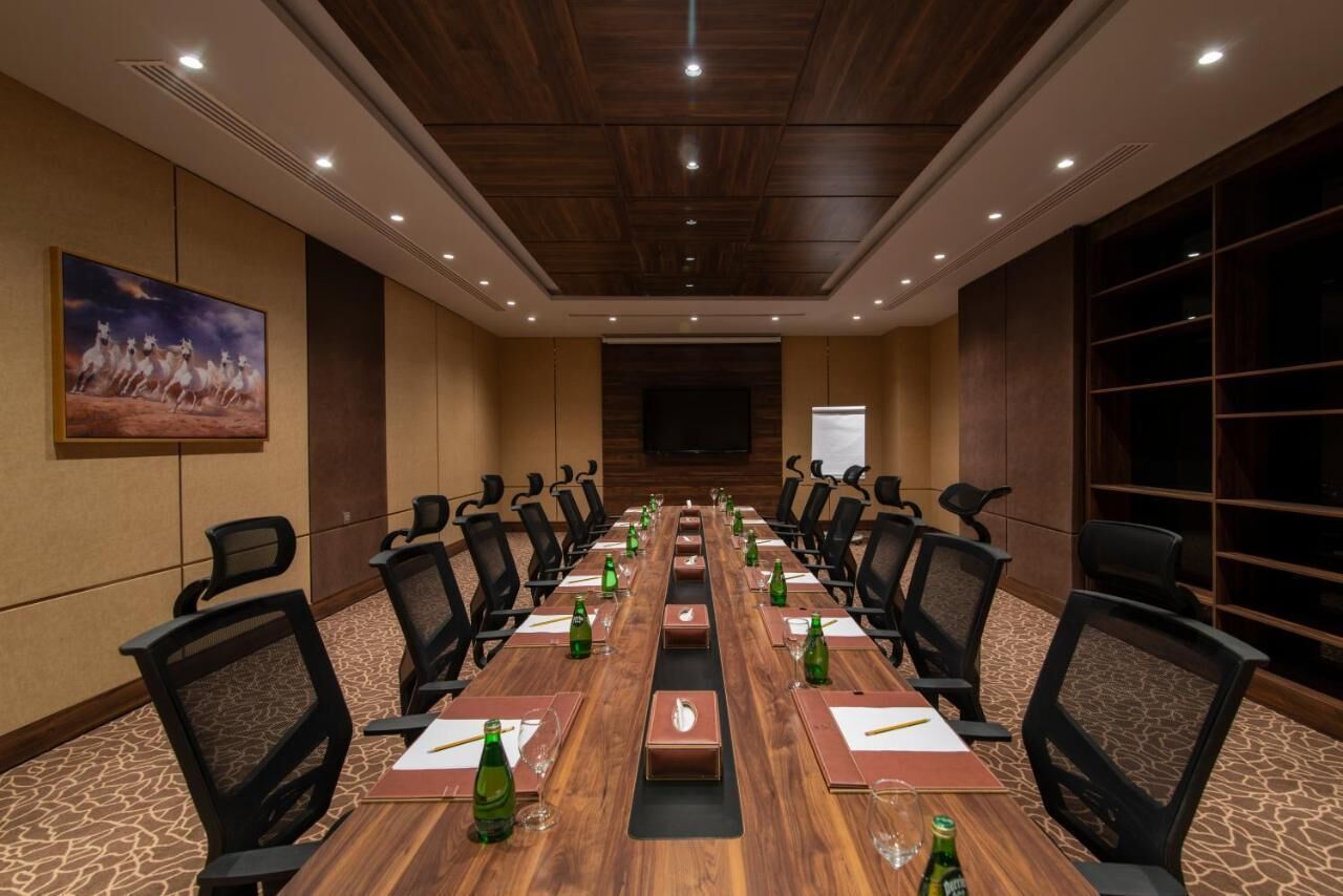 A conference room with a long wooden table, black chairs, and a projector screen at the front.