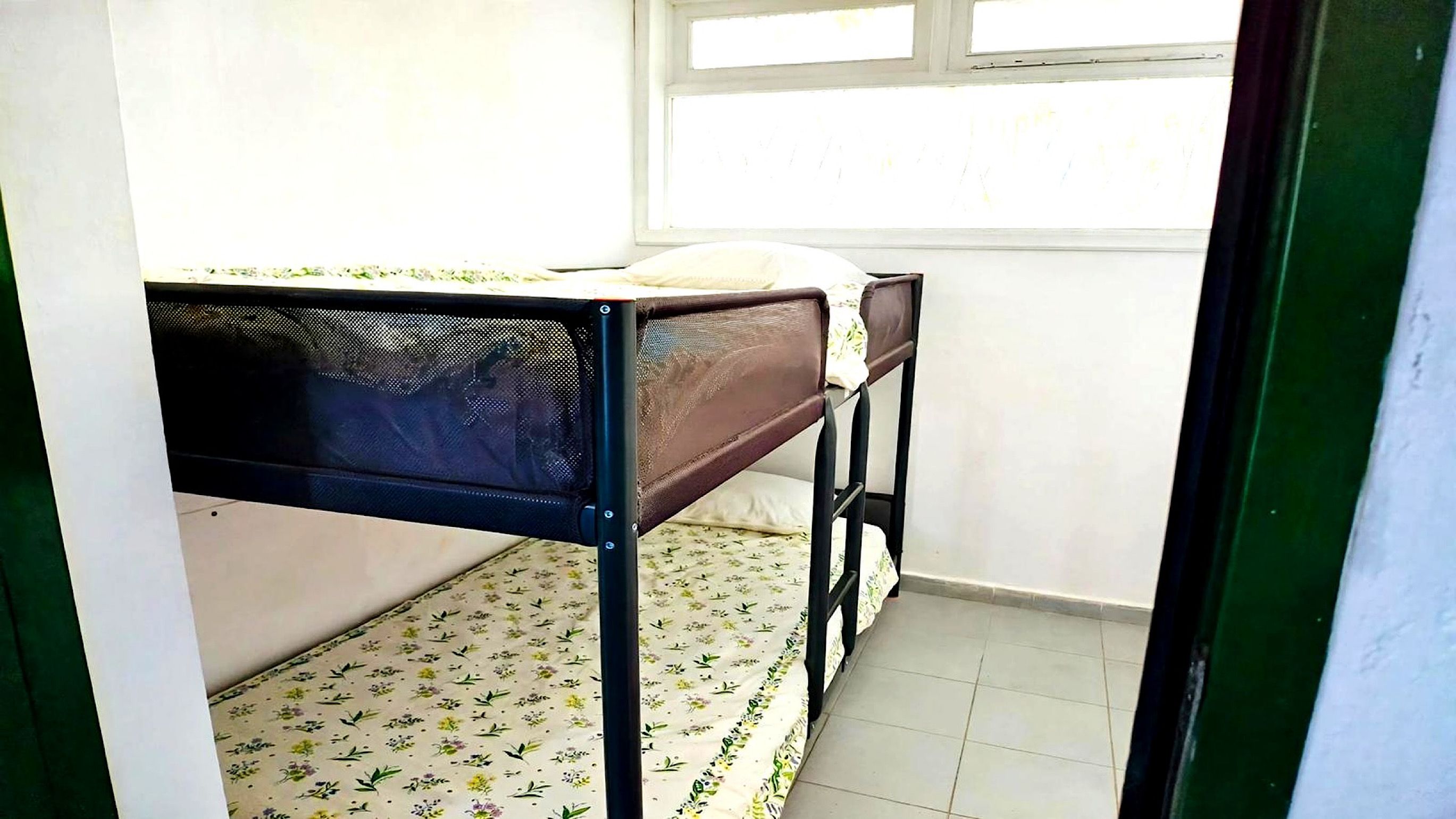 A small room with a single bed, a window, and a tiled floor.