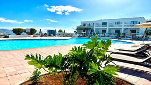 Una piscina con sillas de descanso y sombrillas, rodeada por un edificio de fachada blanca y vistas al mar.