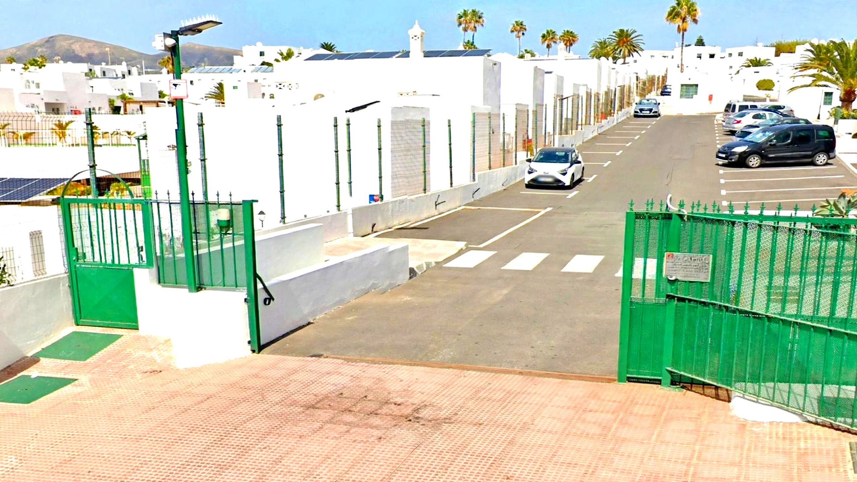 A street view with a gated entrance, parked cars, and buildings with white facades.