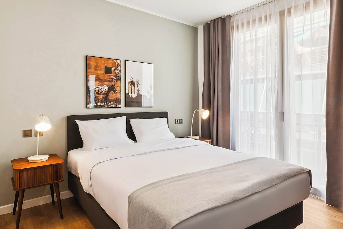 Moderni hotellihuone, jossa on suuri sänky, kaksi yöpöytää valaisimineen, suuri ikkuna läpikuultavine verhoineen sekä kaksi seinälle asennettua taideteosta.