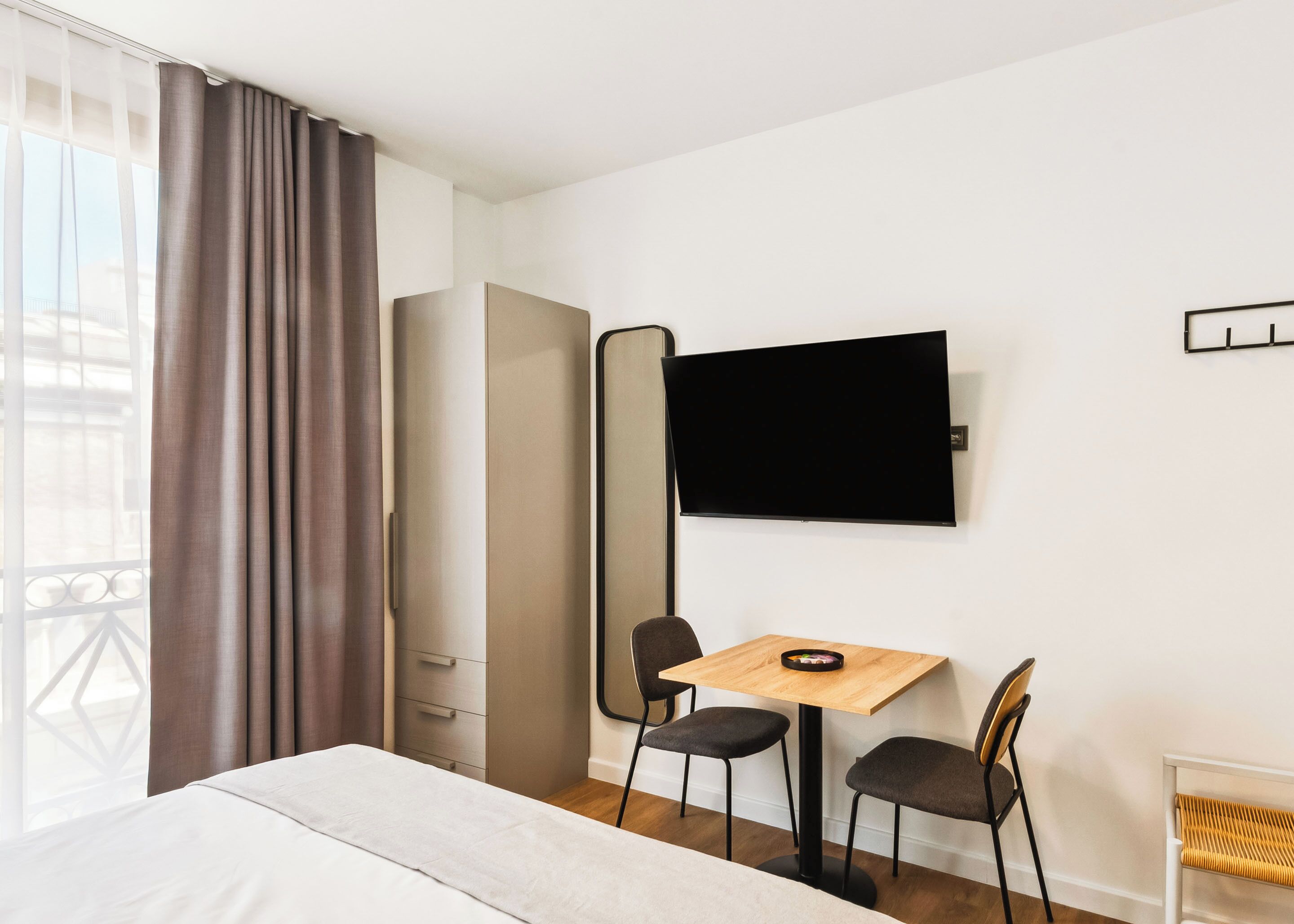 A hotel room with a bed, a desk with two chairs, a television on the wall, and a window with curtains.