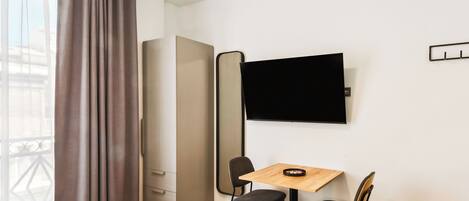 A hotel room with a bed, a desk with two chairs, a television on the wall, and a window with curtains.