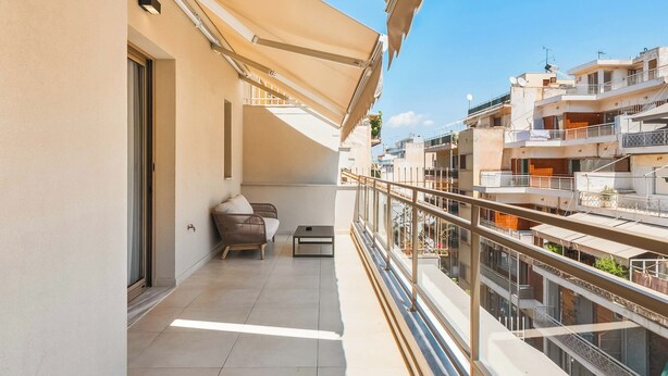 A balcony with a sofa and a table, a glass railing, and a view of neighboring buildings. - Limehome Piraeus Port (Piraeus)