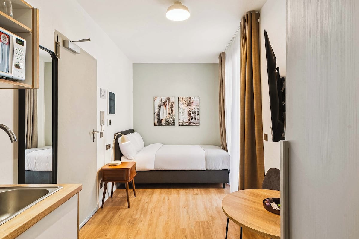 Kompakti hotellihuone, jossa on sänky, yöpöytä ja pieni keittonurkkaus.
