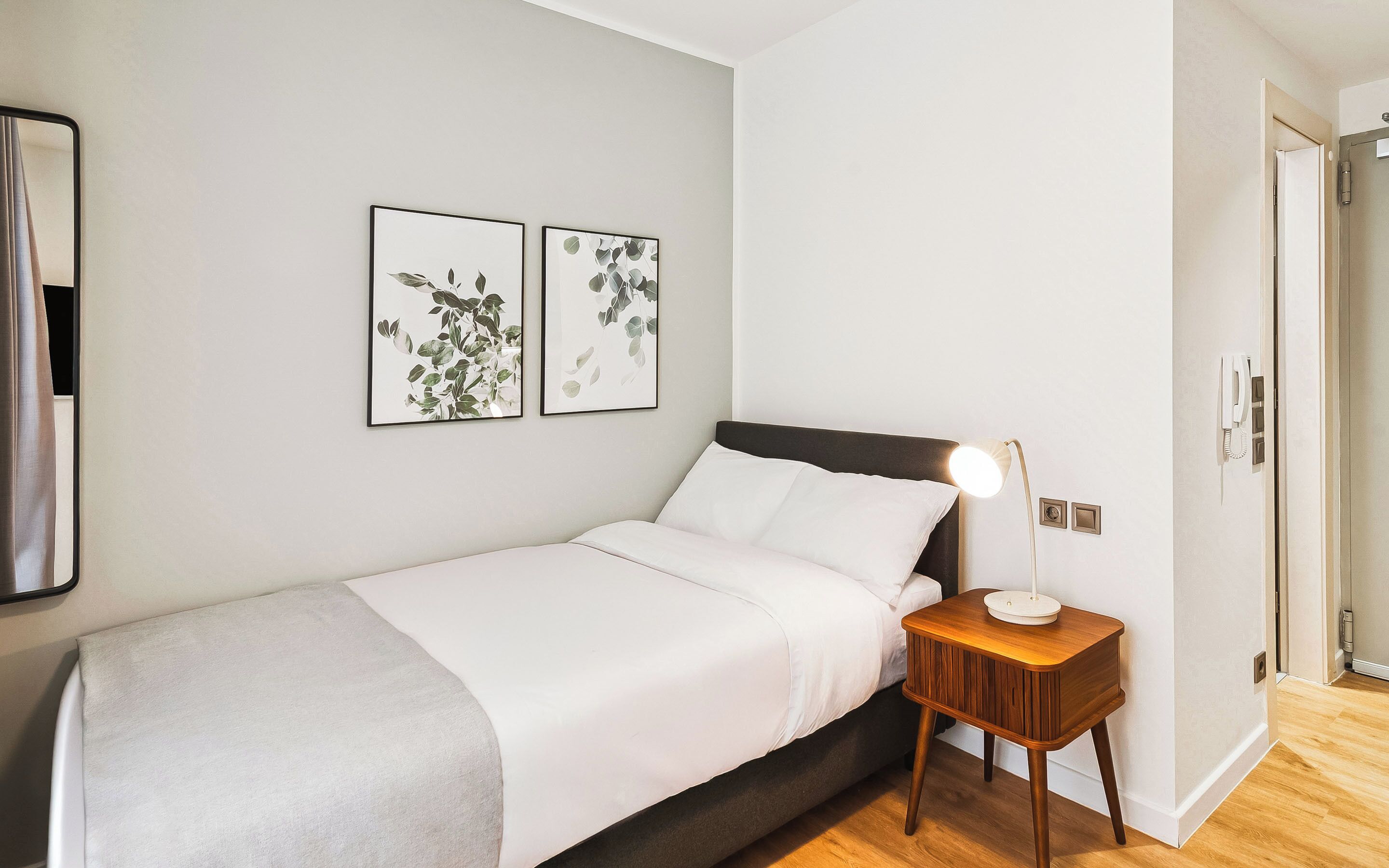 A neatly made bed with a white comforter and pillows, a bedside table with a lamp, and two framed botanical prints on the wall.