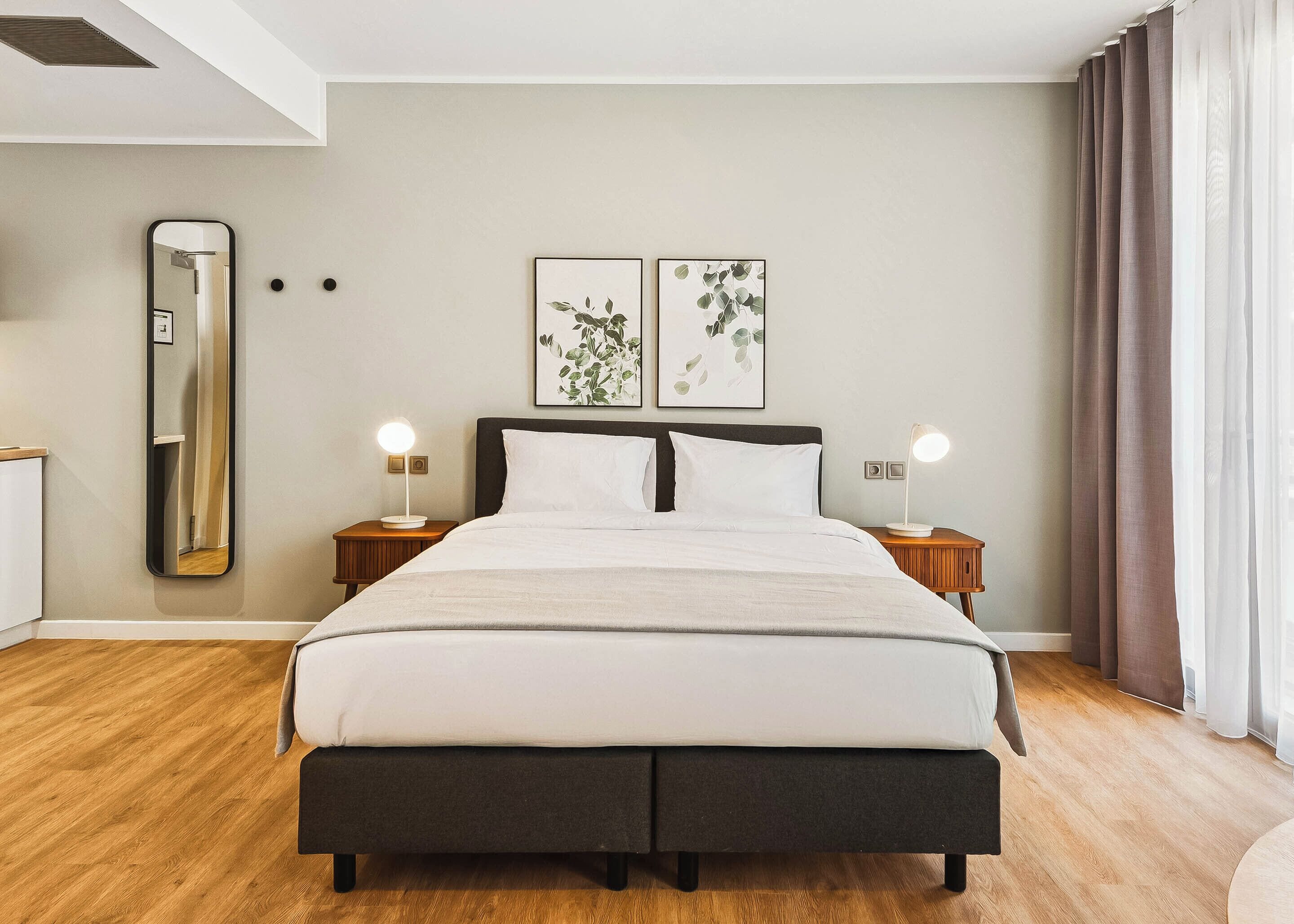Une chambre moderne avec un grand lit, deux tables de chevet avec des lampes, un miroir fixé au mur et deux reproductions botaniques encadrées.
