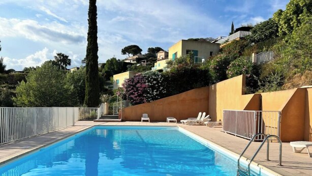 A rectangular swimming pool with clear blue water, surrounded by a tiled deck. There are lounge chairs and a metal ladder for access. The pool area is enclosed by a low fence, and there are buildings and trees in the background. - Quiet house on the Côte d'Azur (Cagnes-sur-Mer)