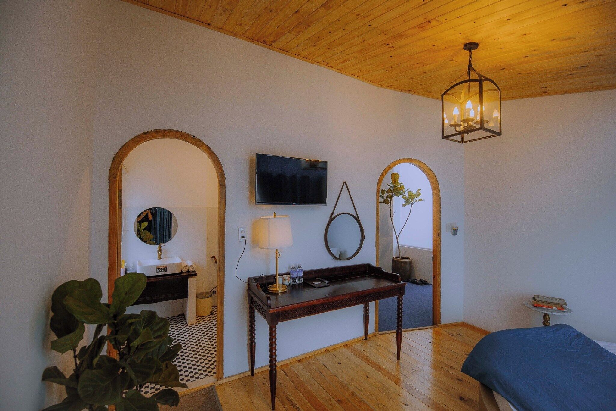 A bedroom with a wooden ceiling, a bed, a bedside table with a lamp, a TV mounted on the wall, and a mirror.