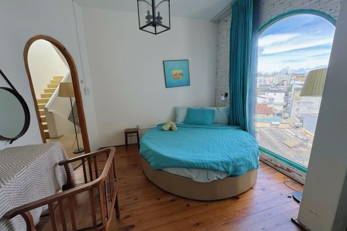 Une chambre avec un lit rond, une chaise en bois, un miroir et une vue sur la ville par une fenêtre.