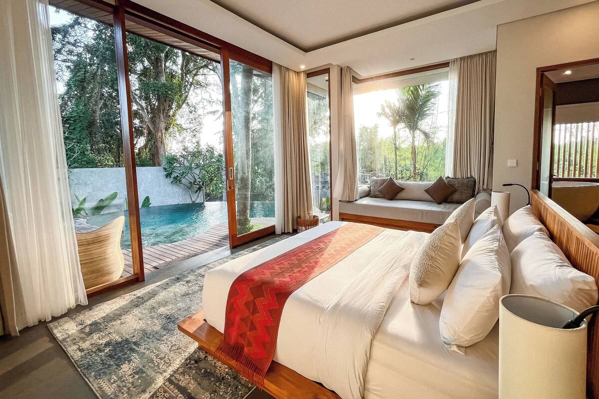 A spacious bedroom with a large bed, a view of a swimming pool, and a balcony with a sofa.