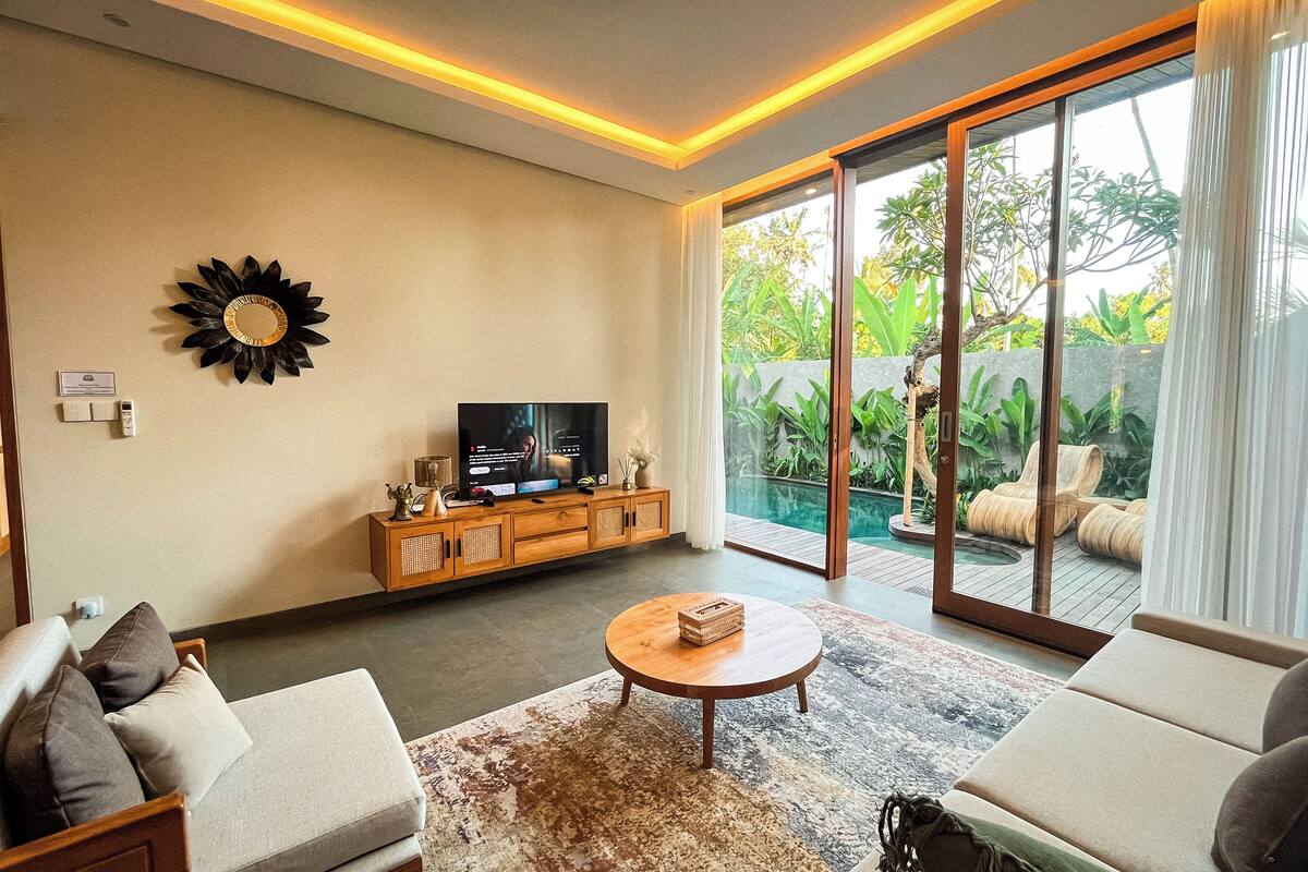 A modern living room with a flat-screen TV, wooden cabinet, and a large sliding glass door leading to a pool area.