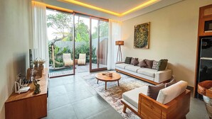 A modern living room with a sofa, coffee table, and a large sliding glass door leading to a garden. - Villa Suarga Des Anicca (Ubud)
