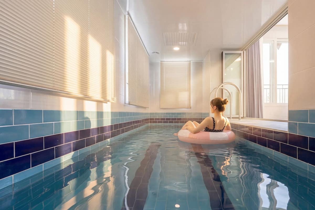 An indoor swimming pool with tiled walls, a person on a float, and windows with blinds.