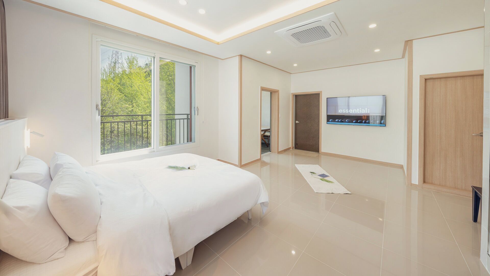 A modern hotel room with a large bed, a TV on the wall, and a balcony with a view of trees.