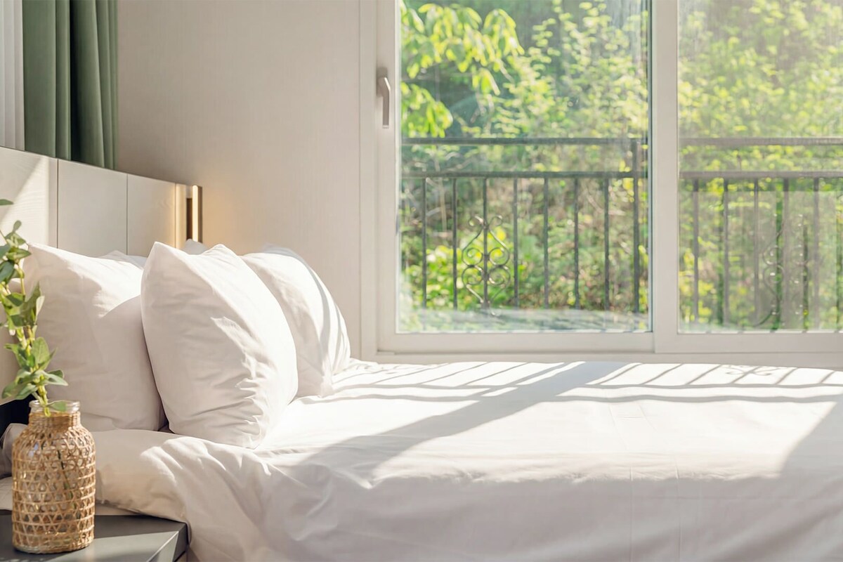 A bedroom with a large window, a bed with white bedding, and a decorative bottle on the nightstand.
