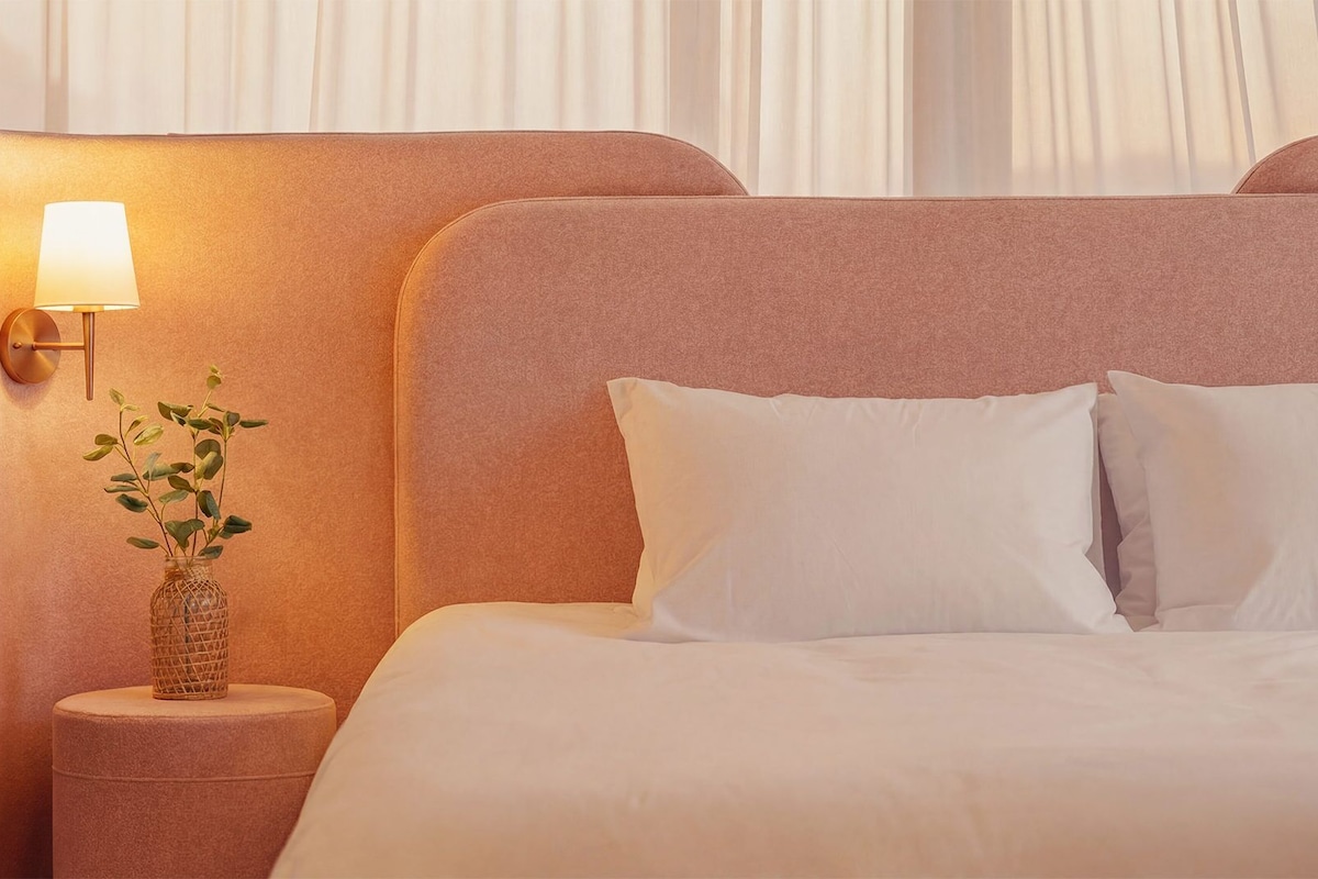 A hotel room with a large bed, white bedding, a nightstand with a lamp, and a small plant.