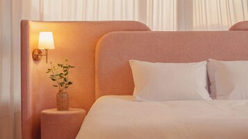 A hotel room with a large bed, white bedding, a nightstand with a lamp, and a small plant.