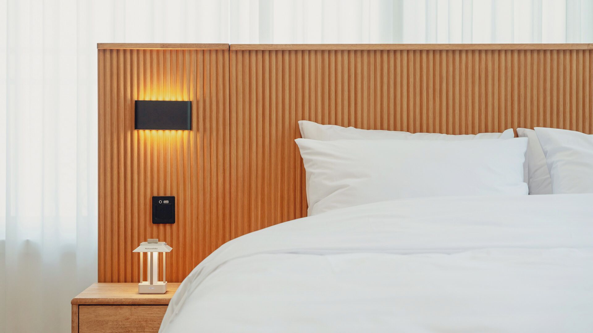 Una habitación de hotel con cabecera de madera, una mesita de noche con lámpara y una cama con ropa blanca.