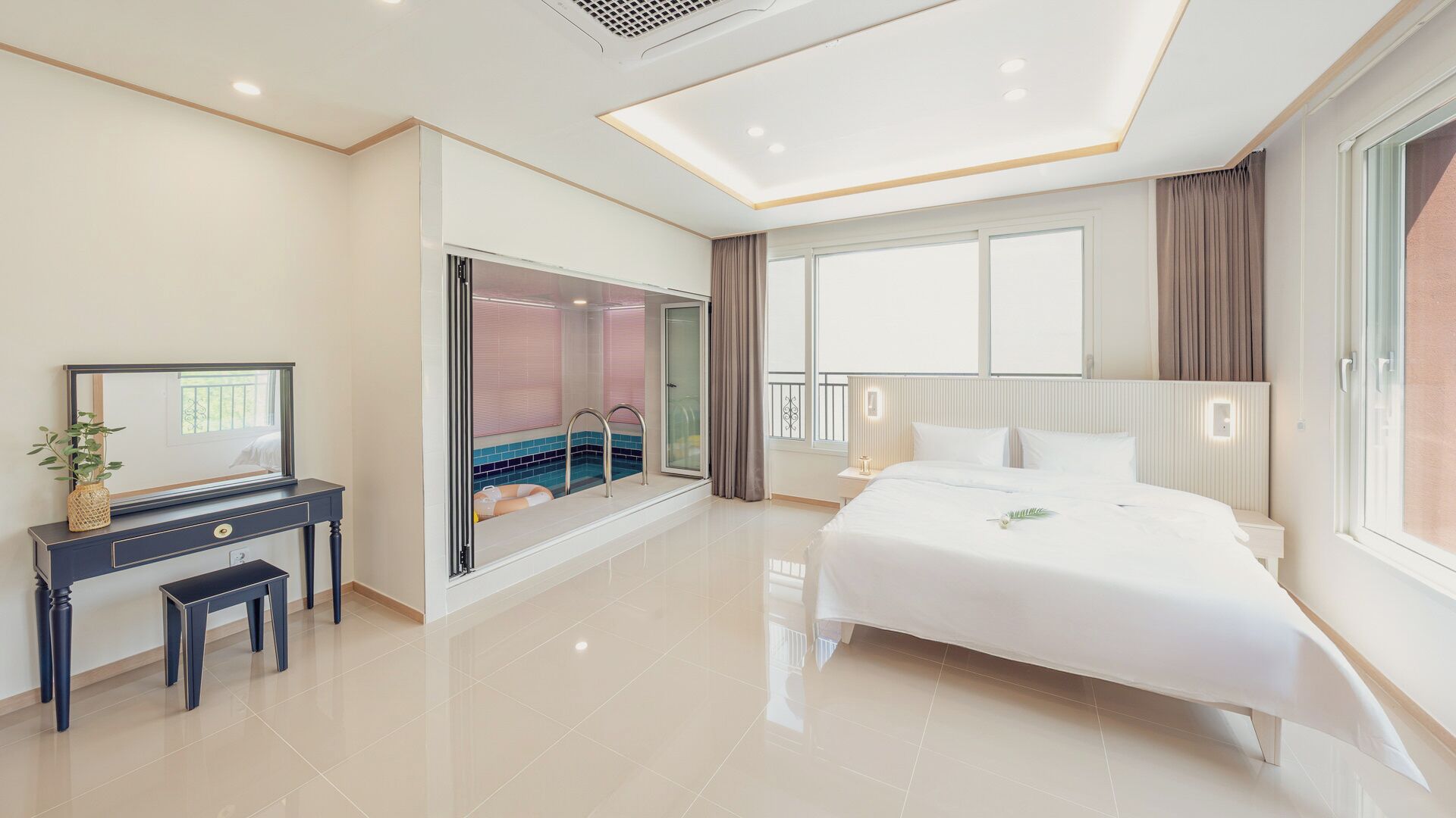 A modern hotel room with a large bed, a desk with a mirror, a sliding glass door to a bathroom, and a large window.