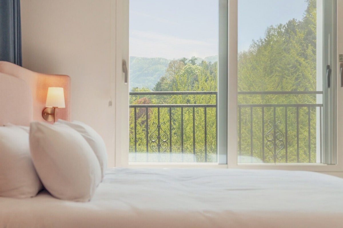 A bedroom with a bed, pillows, a nightstand with a lamp, and a balcony with a view of trees.