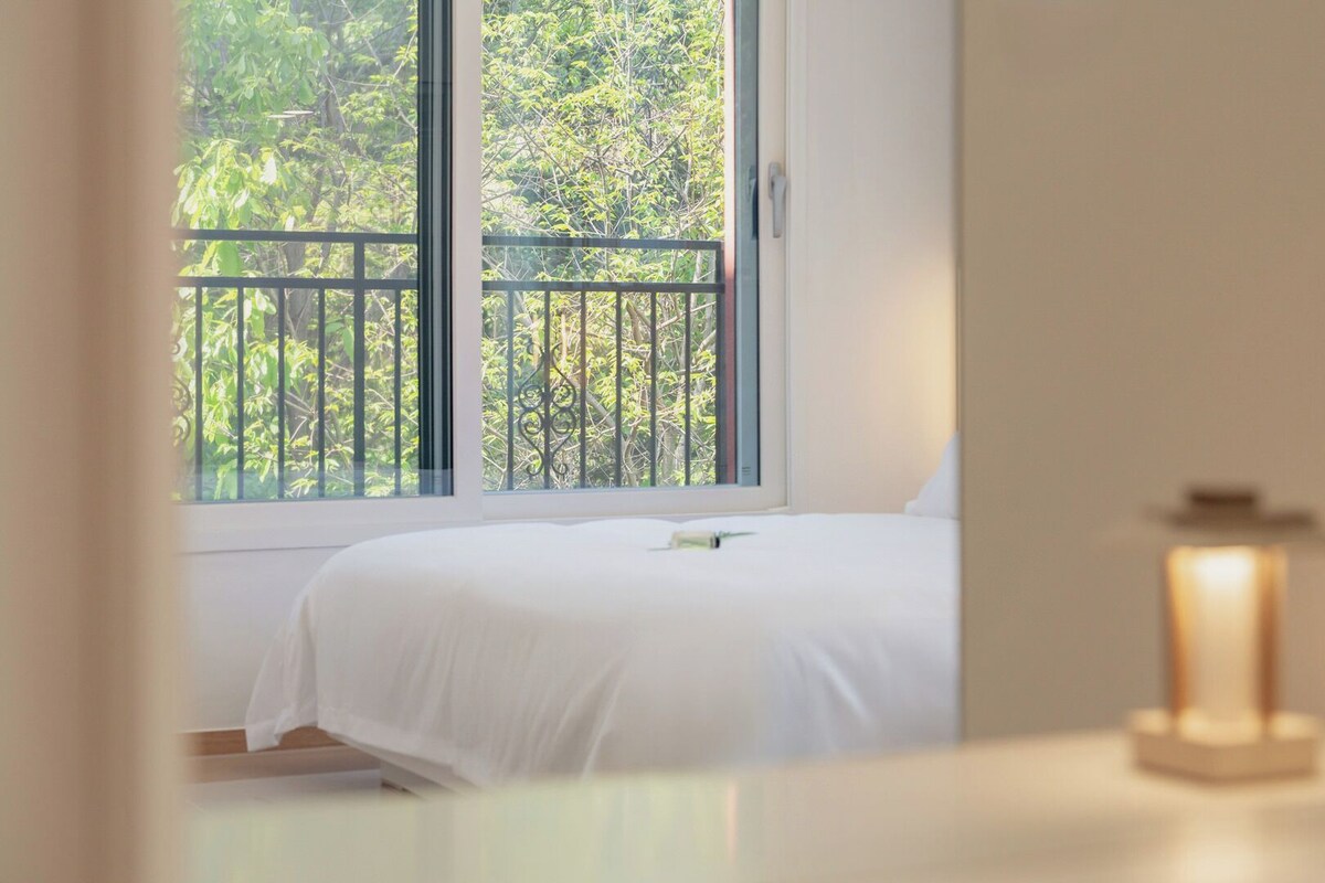A bedroom with a bed, a window with a view of trees, and a bedside table with a lamp.