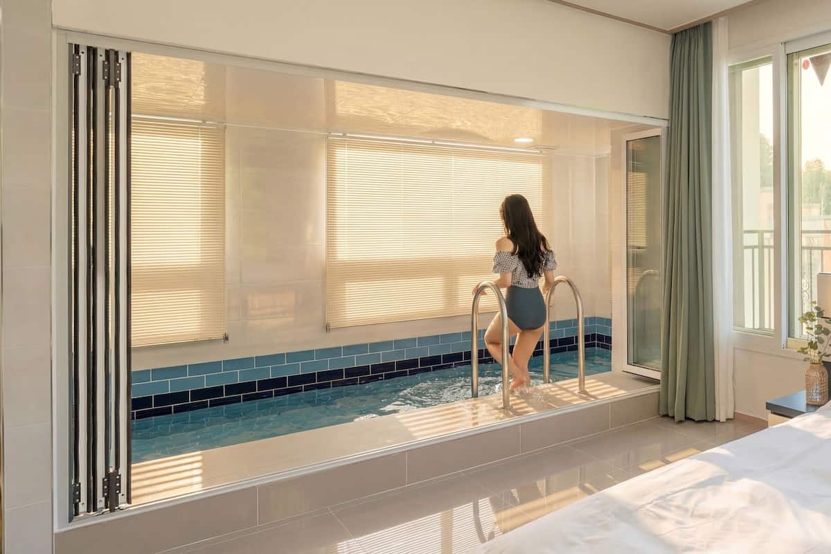 A hotel room with a large window, a pool, and a bed.